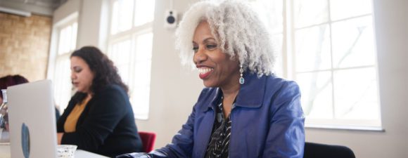 Woman with white curly hair and a blue blazer smiling as she uses a learning management platform on a white laptop computer.