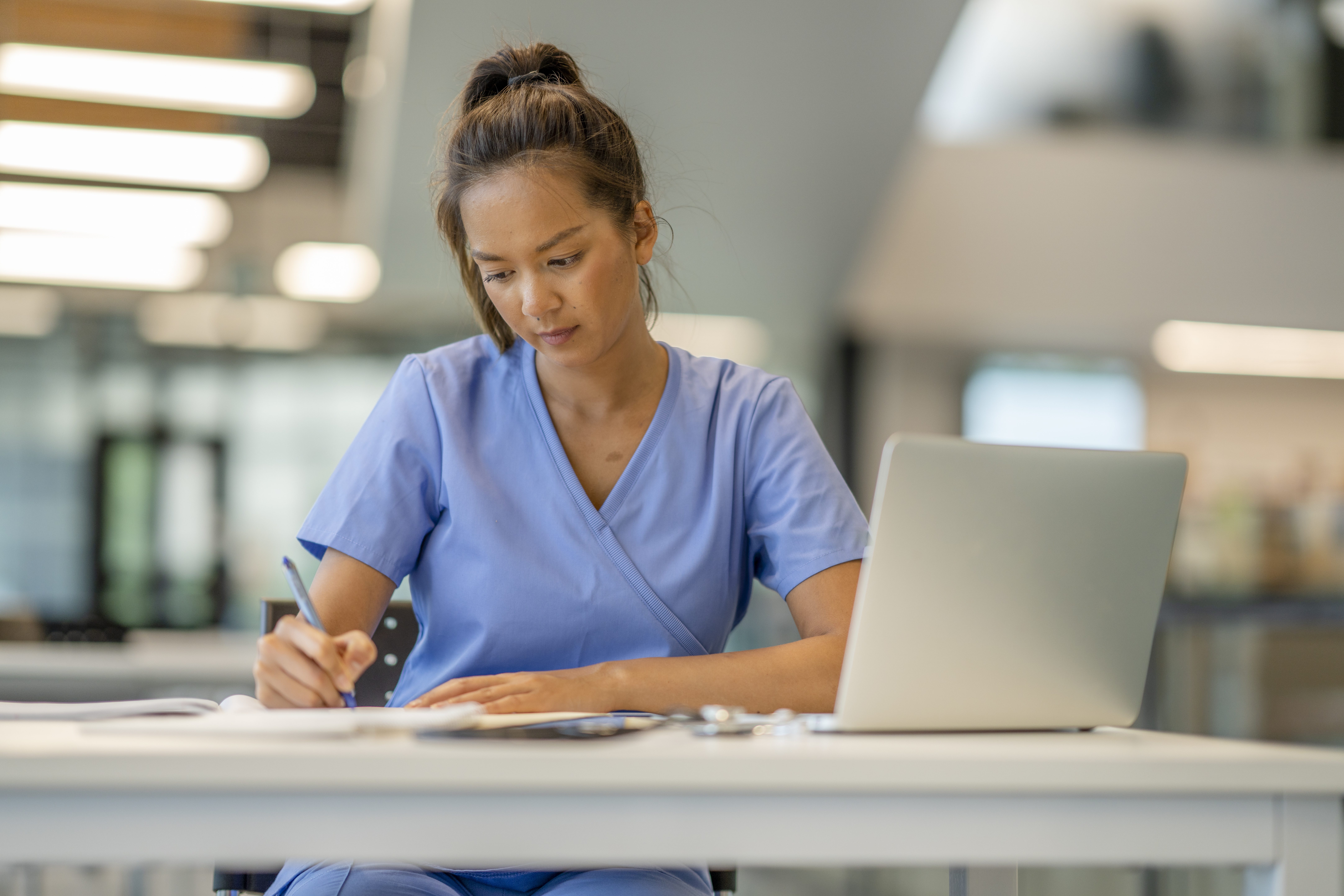 A woman wearing scrubs diligently studies at a desk in a modern educational building.