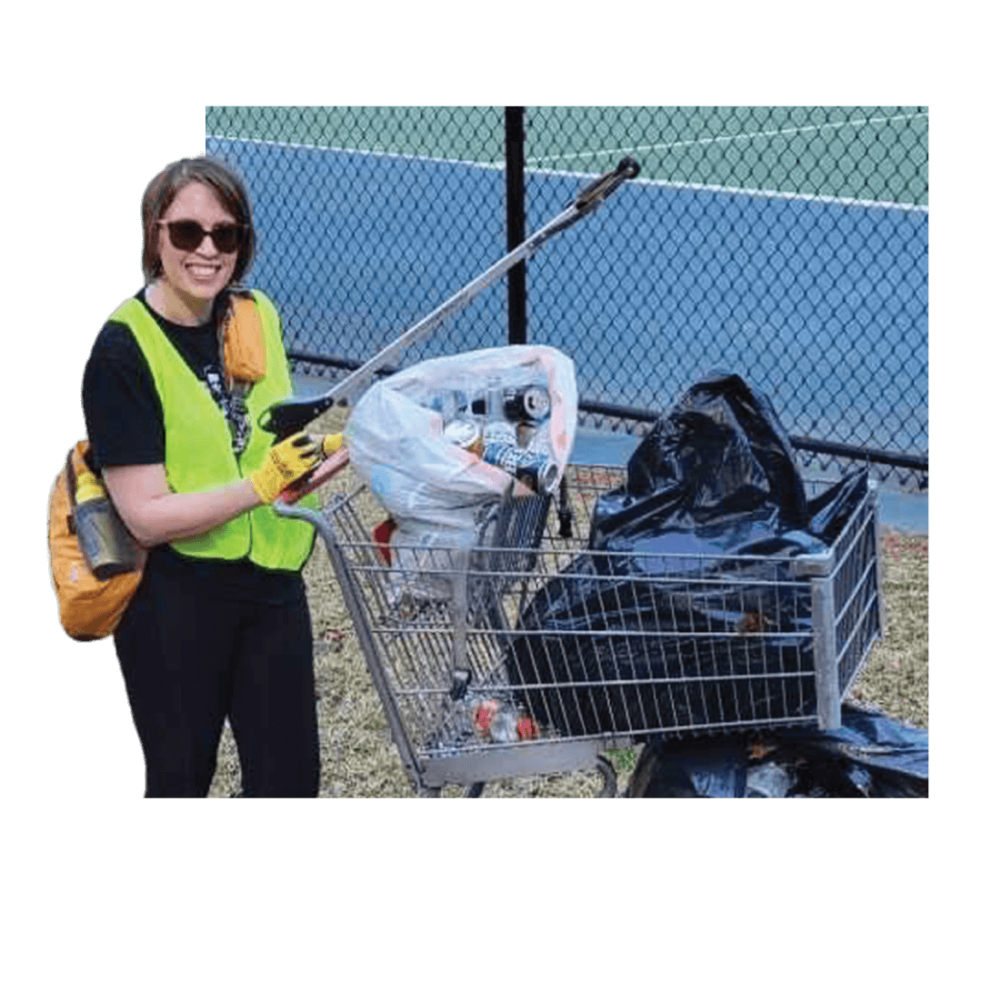 An employee picks up trash outside near a tennis court and put the trash in a bag she is pushing in a shopping cart.