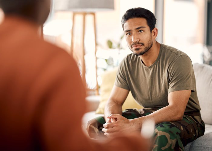 A veteran wearing camo on a couch, discussing mental health with a counselor
